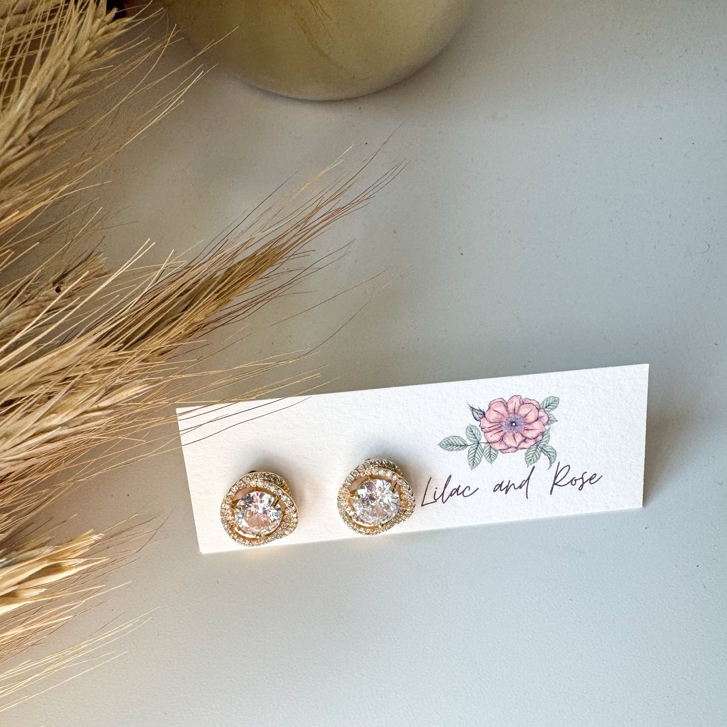 Gold earrings on a card with floral design and 'Lilac and Rose' text, placed on a light surface.