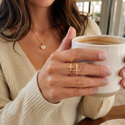 A close-up lifestyle shot of a woman holding a white ceramic coffee mug, wearing a delicate gold ring with two small cross motifs on her ring finger.
