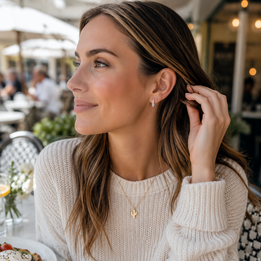 A woman with long brown hair sits at an outdoor cafe table wearing a cream knit sweater and a delicate gold necklace with a small letter pendant. A plate of avocado toast is visible on the table in the foreground.