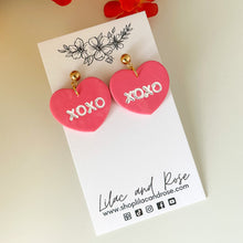 Load image into Gallery viewer, A pair of pink heart-shaped earrings with 'XOXO' printed in white on each heart, displayed on a white card with floral patterns and the brand name 'Lilac and Rose'.