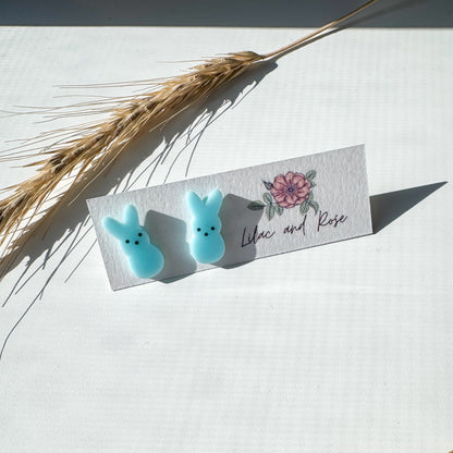 Two blue bunny-shaped earrings on a card with floral design and text, placed on a light surface with dried plants.