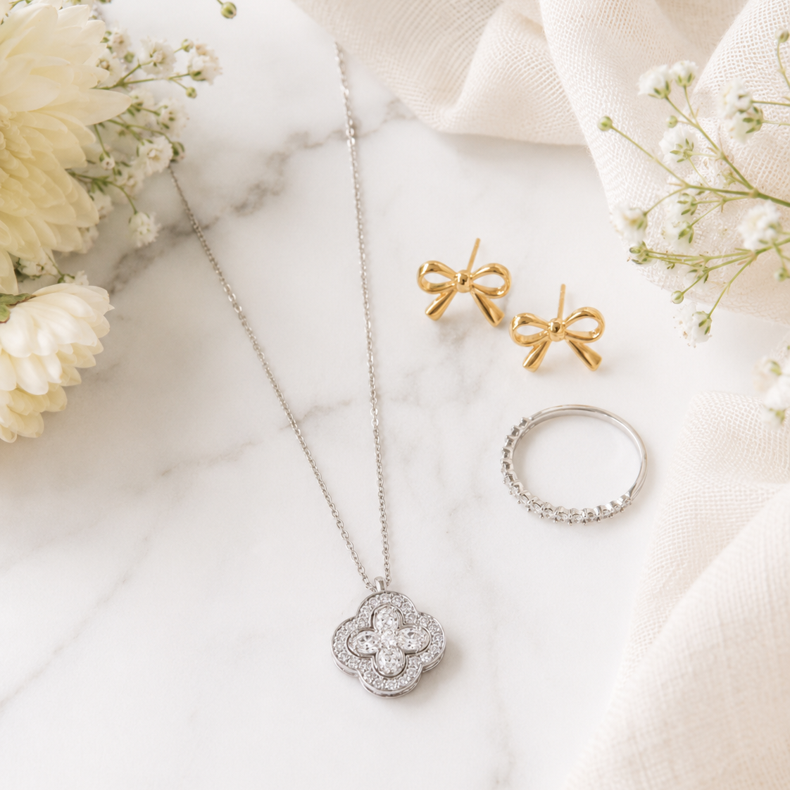 Silver and gold jewelry set featuring a clover pendant necklace, bow stud earrings, and a ring, styled on a white marble surface with cream fabric and baby's breath flowers.