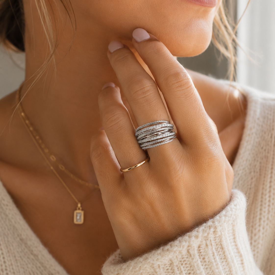 A close-up lifestyle shot of a woman wearing a stack of silver rings on her finger, along with a delicate gold necklace and a cream knit sweater.