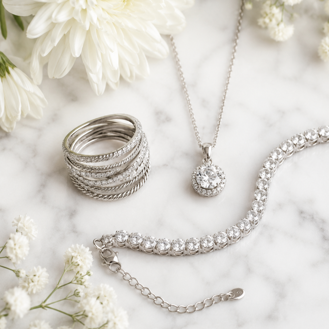 Silver jewelry set including a tennis bracelet, a pendant necklace, and a stacked ring, styled on a white marble surface with white flowers.