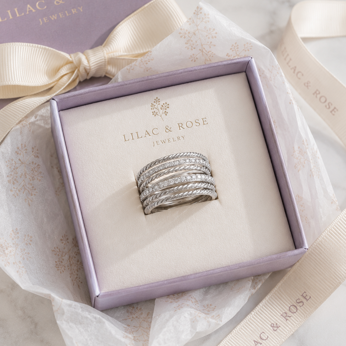 A silver ring set with clear pavé stones displayed in a lavender Lilac & Rose jewelry box, surrounded by cream ribbon and tissue paper on a marble surface.