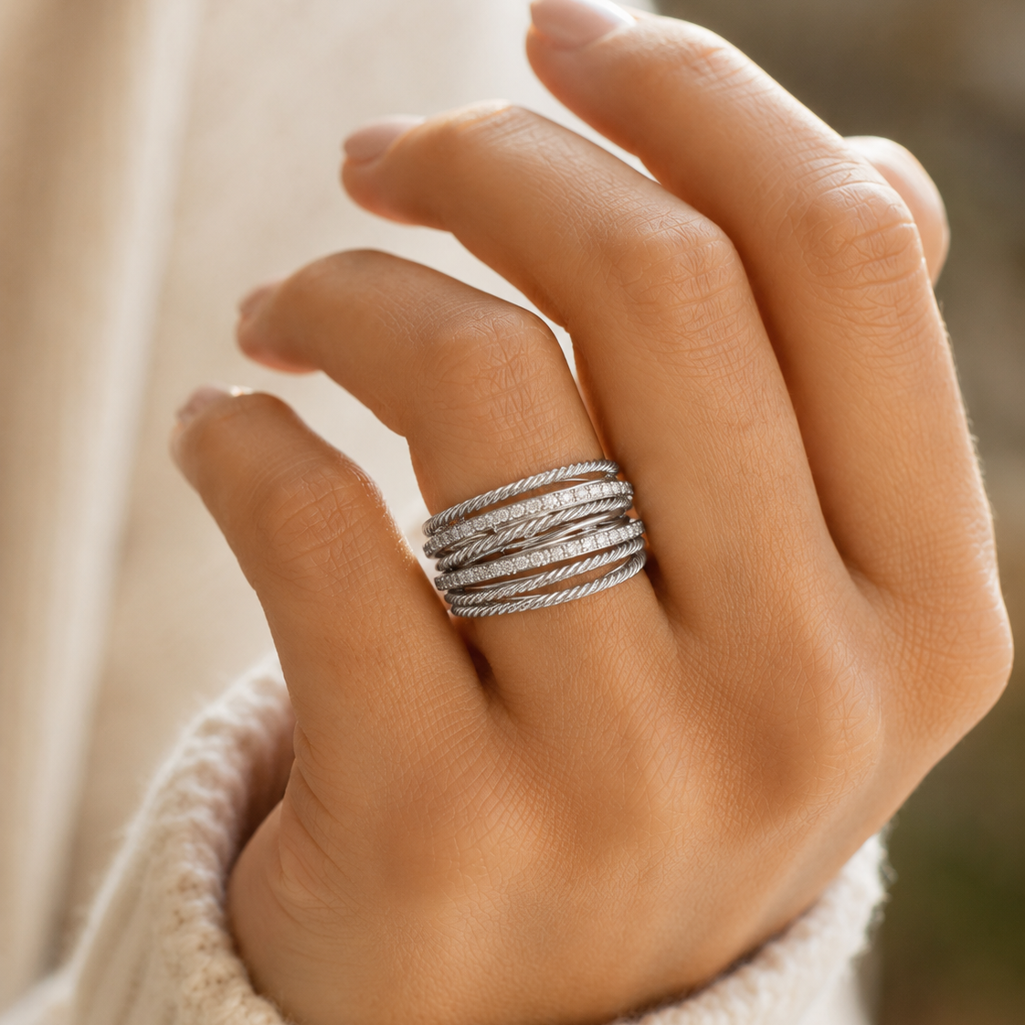 A close-up of a hand wearing a stack of textured silver rings, styled with a cream knit sweater against a soft, blurred background.