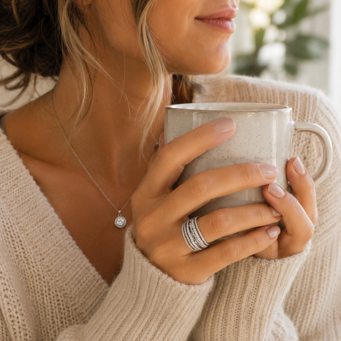 A close-up lifestyle shot of a woman in a beige knit sweater holding a ceramic mug, wearing a delicate silver necklace with a round clear stone and stacked silver rings on one finger.