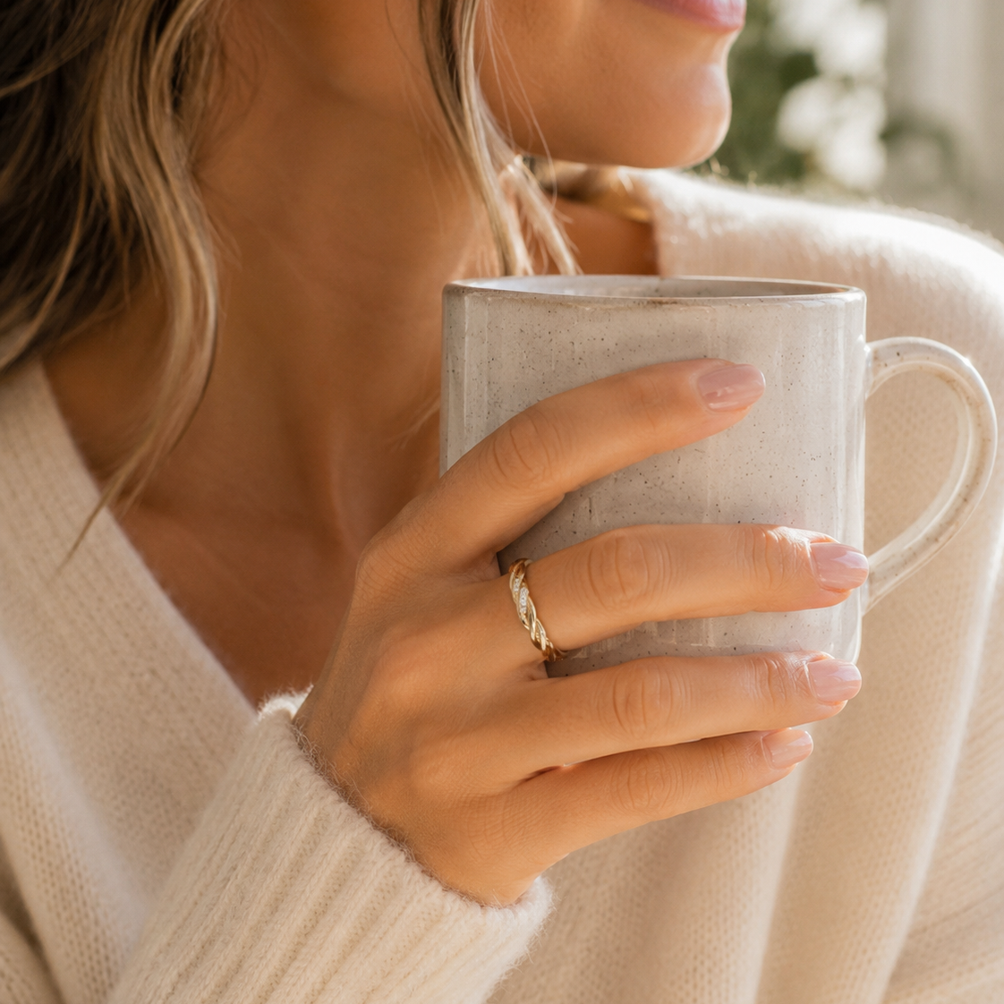 A close-up lifestyle shot of a woman in a cream knit cardigan holding a textured light gray ceramic mug, wearing a delicate gold band ring on her finger.