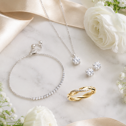 A styled flat-lay of silver jewelry including a necklace, bracelet, and stud earrings, alongside a gold ring, arranged on a white marble surface with white roses and cream satin ribbons.