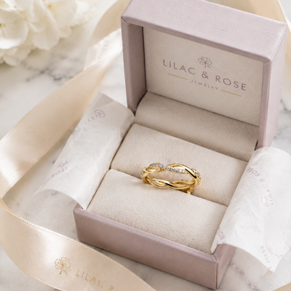 A gold ring with a small clear gemstone set in a lavender Lilac & Rose jewelry box, styled on a marble surface with a cream ribbon and white flowers.