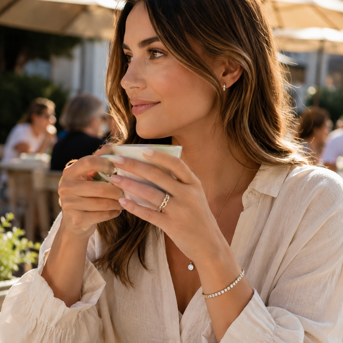 A woman in a white blouse at an outdoor cafe, holding a cup and wearing delicate silver jewelry including a ring, bracelet, and small pendant necklace.