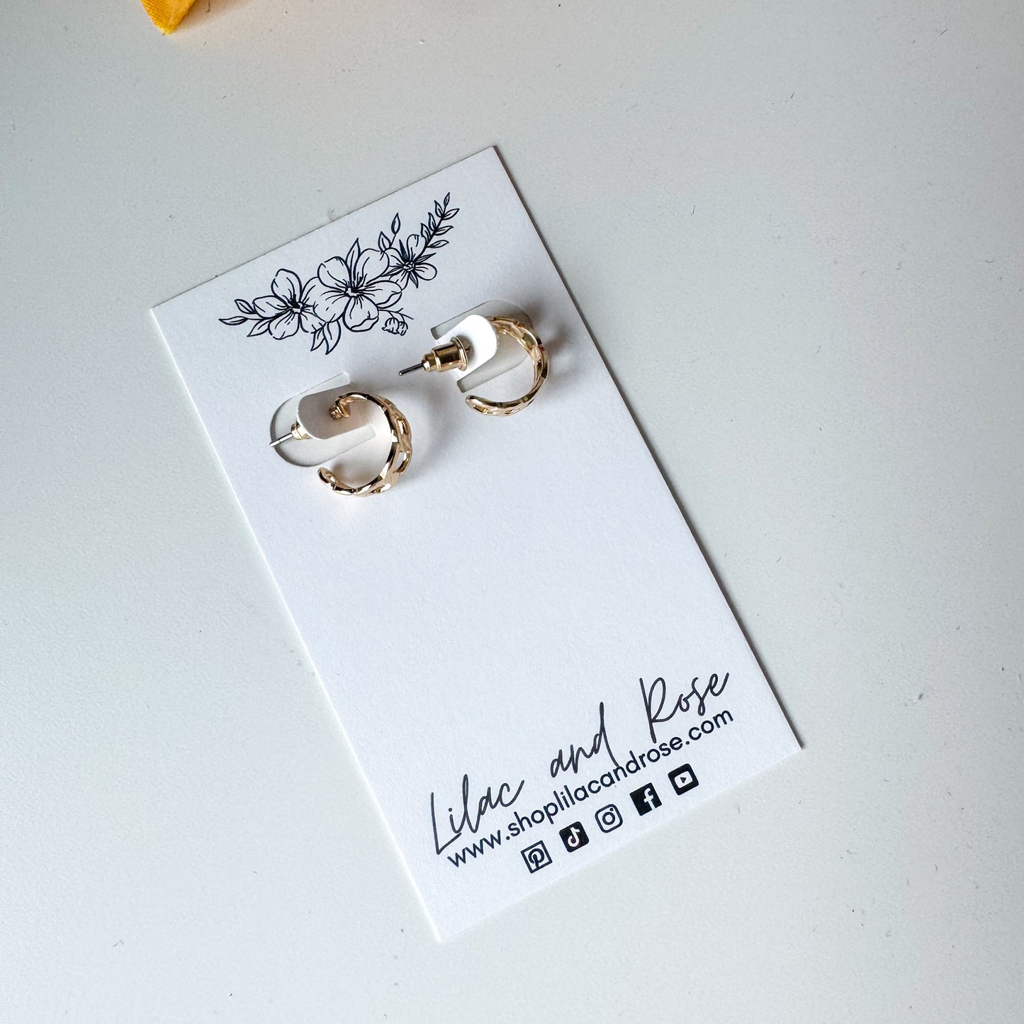 A pair of silver mini chain hoop earrings displayed on a white card with floral pattern and the brand name 'Lilac and Rose' written at the bottom.