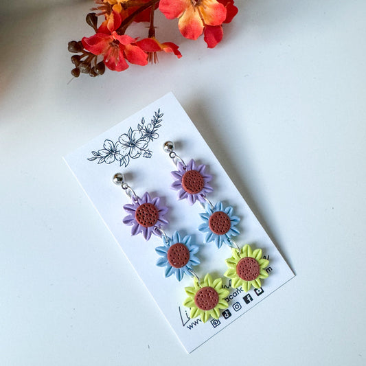 A set of three handcrafted earrings made from polymer clay, designed to look like flowers in vibrant colors, displayed on a white card.