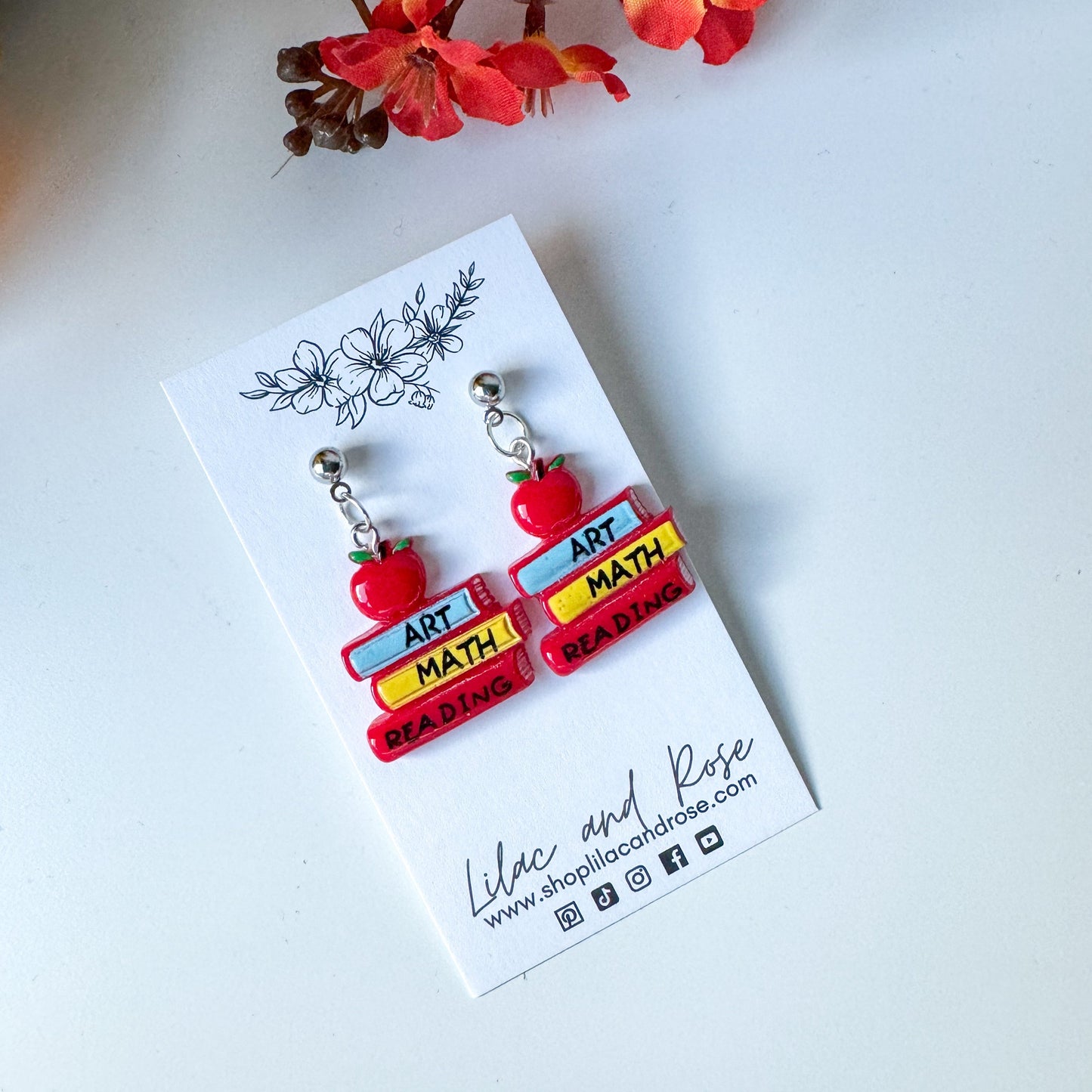 A pair of earrings designed to look like red and yellow school books with the labels 'ART MATH P.E.' on a white card.