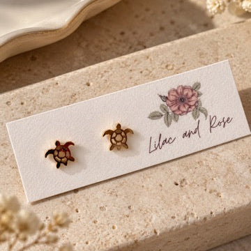 Two small turtle-shaped stud earrings displayed on a white card labeled 'Lilac and Rose,' placed on a beige stone surface with dried flowers and a ceramic dish in the background.