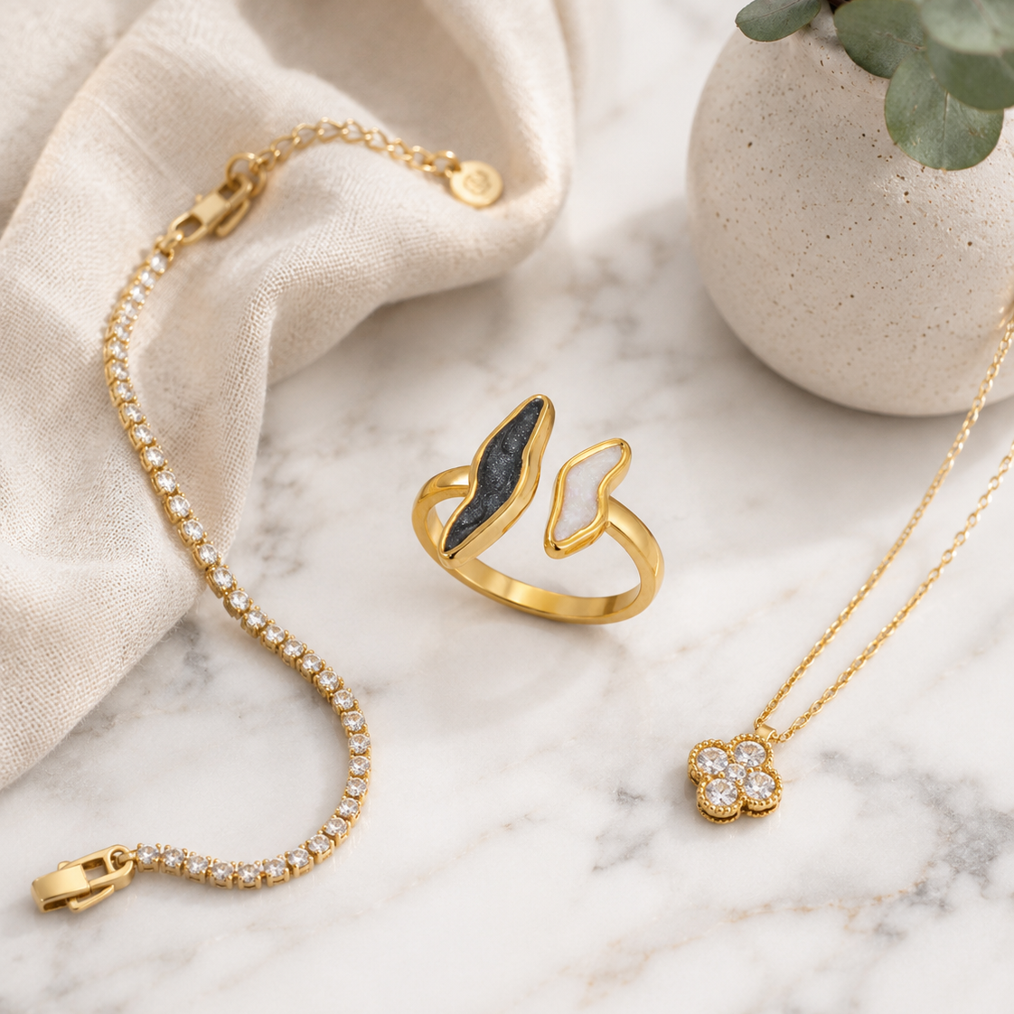 Gold jewelry set including a ring with a black inlay, a delicate chain bracelet, and a flower-shaped pendant necklace, styled on a marble surface with a beige fabric drape and a small potted eucalyptus plant.