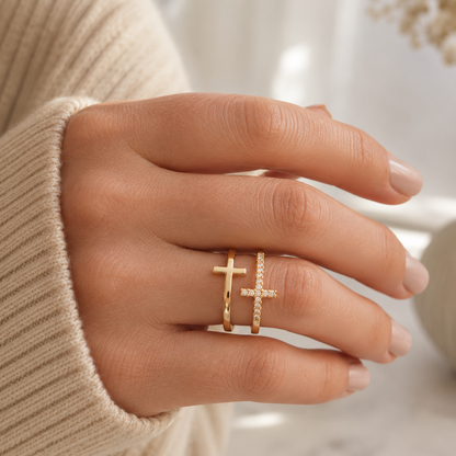 A close-up of a hand wearing two gold rings: one with a small cross and the other with a pavé-set cross, styled with a beige ribbed sweater sleeve and a soft-focus decorative vase in the background.
