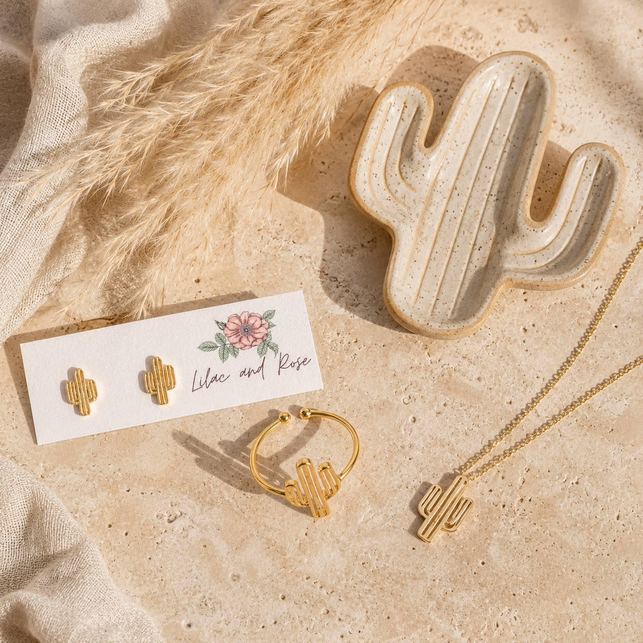 Gold cactus-themed jewelry set including a necklace, hoop, and stud earrings on a white card, styled on a beige stone surface with a ceramic cactus dish and dried grass.
