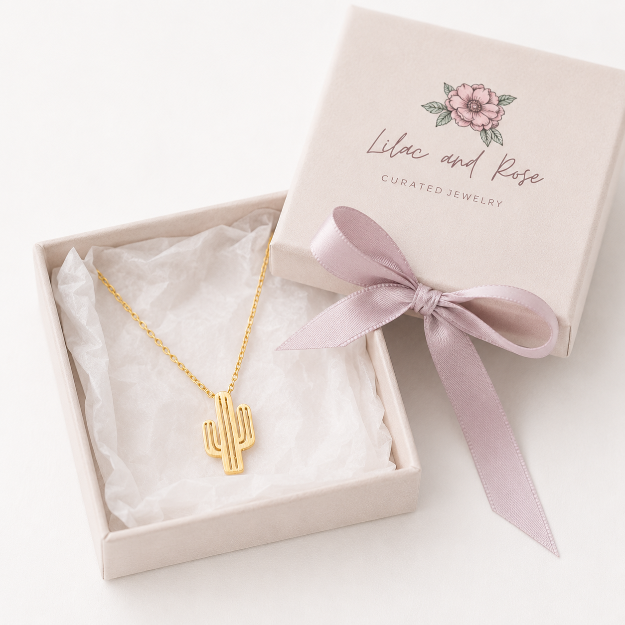 A gold cactus pendant necklace displayed in an open cream jewelry box with white tissue paper, alongside the closed matching box lid printed with the brand name 'Lilac and Rose'.