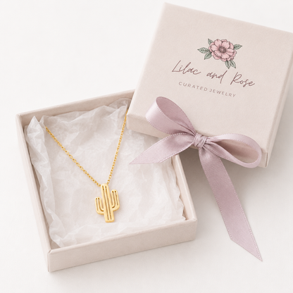 A gold cactus pendant necklace displayed in an open cream jewelry box with white tissue paper, alongside the closed matching box lid printed with the brand name 'Lilac and Rose'.