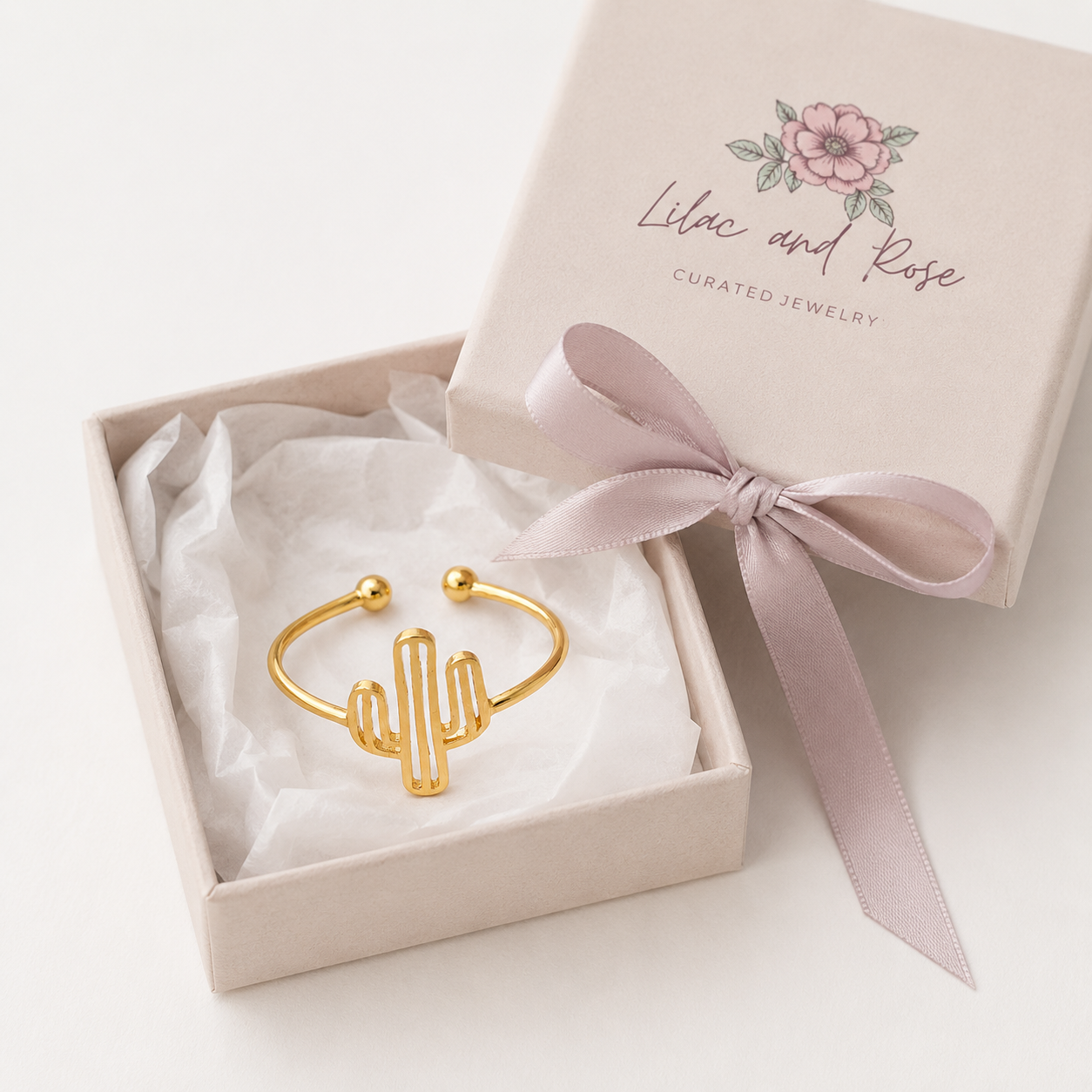 A gold cactus-shaped cuff bracelet presented in an open cream-colored jewelry box with white tissue paper and a dusty pink ribbon. The lid of the box is visible in the background, printed with the brand name 'Lilac and Rose Curated Jewelry' and a pink flower illustration.
