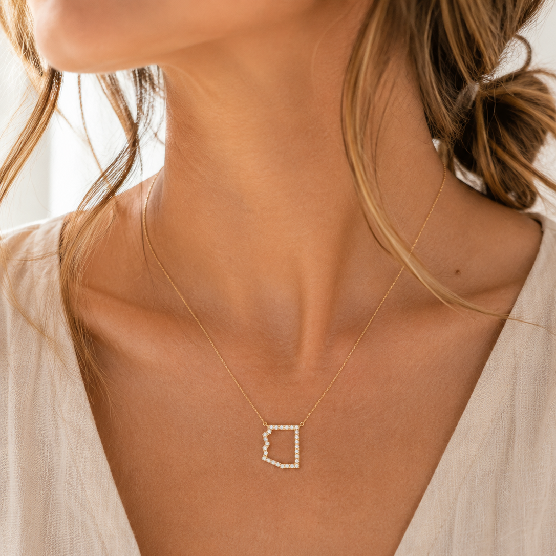 A close-up lifestyle shot of a model wearing a delicate gold necklace with a small, open Arizona pendant studded with tiny clear stones.
