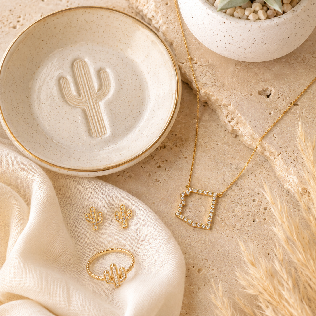 Gold jewelry set featuring an Arizona pendant necklace, a cactus ring, a cactus stud earring, and a matching cactus charm, styled on a beige stone surface with a ceramic dish, fabric, and dried grass.