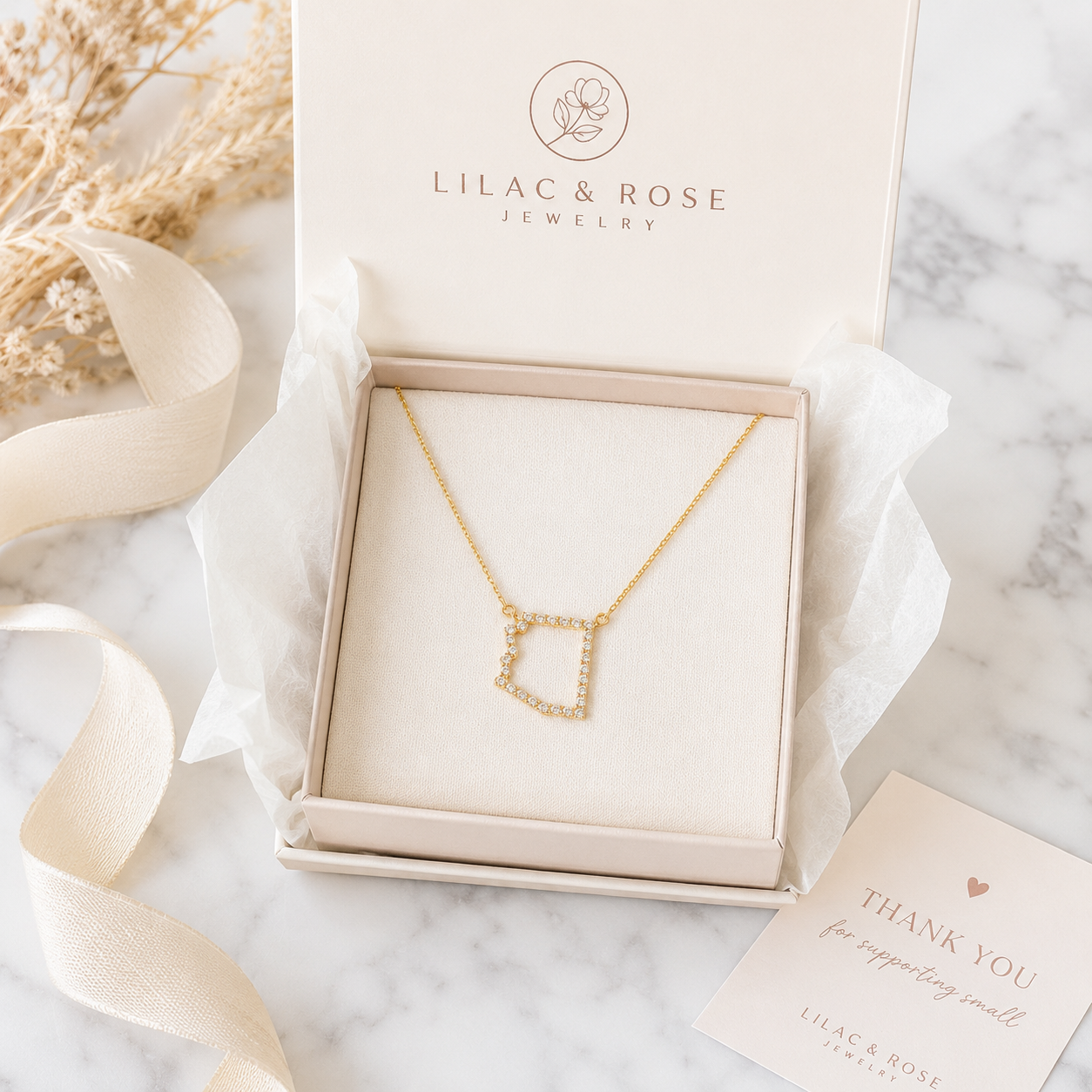 A gold necklace with a geometric Arizona pendant displayed inside an open Lilac & Rose Jewelry gift box on a marble surface, with ribbon, dried flowers, and a thank-you card around it.
