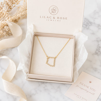 A gold necklace with a geometric Arizona pendant displayed inside an open Lilac & Rose Jewelry gift box on a marble surface, with ribbon, dried flowers, and a thank-you card around it.