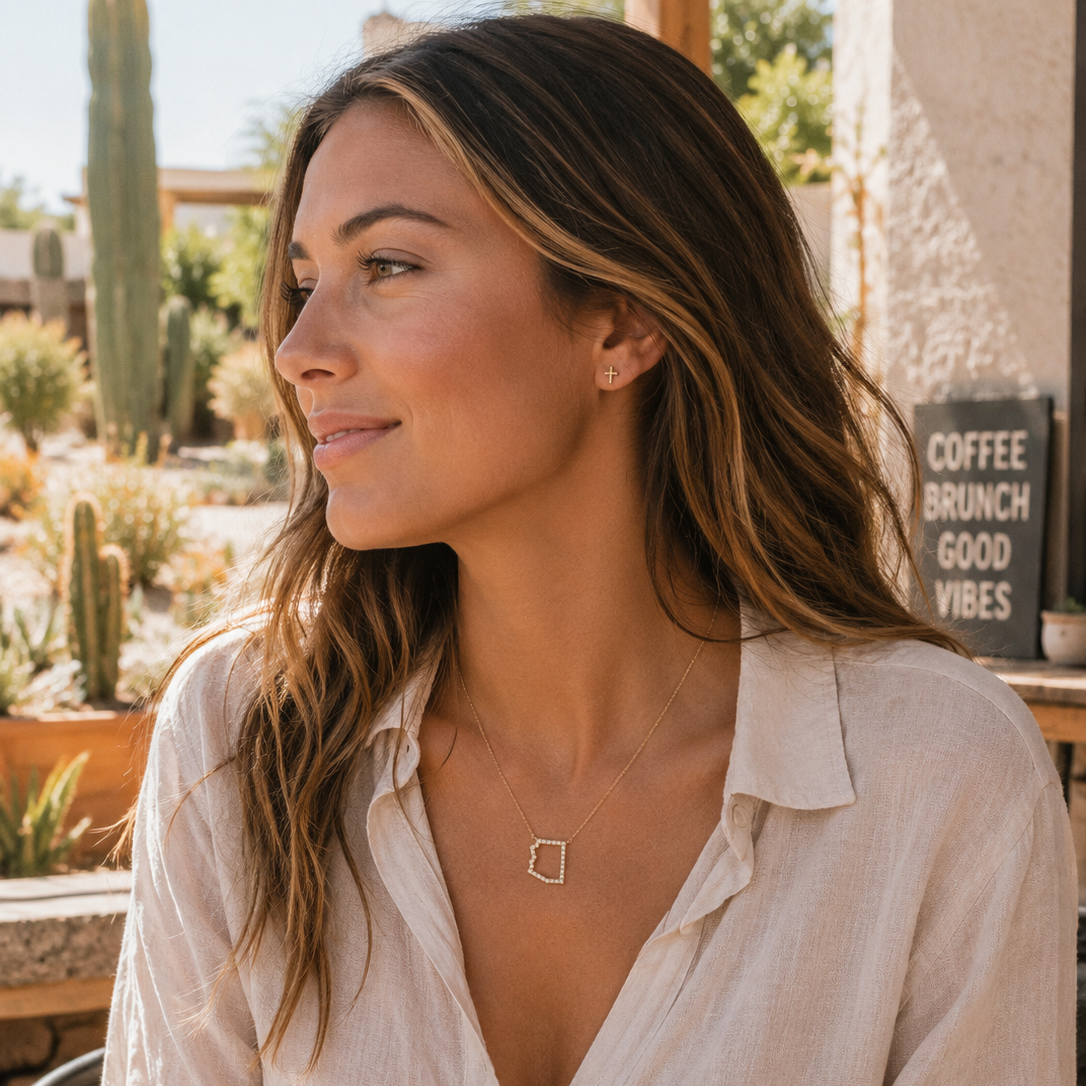 A woman wearing a delicate gold Arizona pendant necklace with an abstract shape, styled with a white linen shirt in a warm, sunny outdoor patio setting.