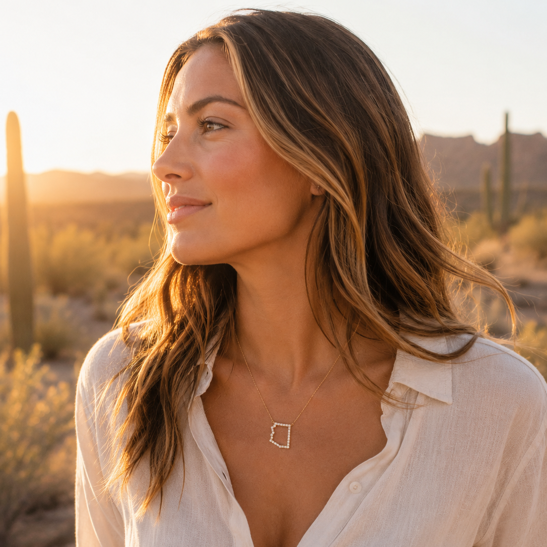 A woman in a desert landscape at sunset wearing a delicate silver necklace with a small open Arizona pendant and a white button-up shirt.