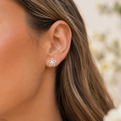 A close-up side profile of a woman's ear wearing a small silver flower-shaped stud earring, with her brown hair and skin visible against a softly blurred light background.