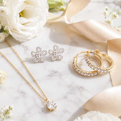 A styled flat-lay of gold and silver jewelry on a white marble surface with white flowers and a satin ribbon. The set includes a gold chain necklace with a clear stone pendant, a pair of silver flower-shaped stud earrings, and a pair of gold hoop earrings with clear stones.