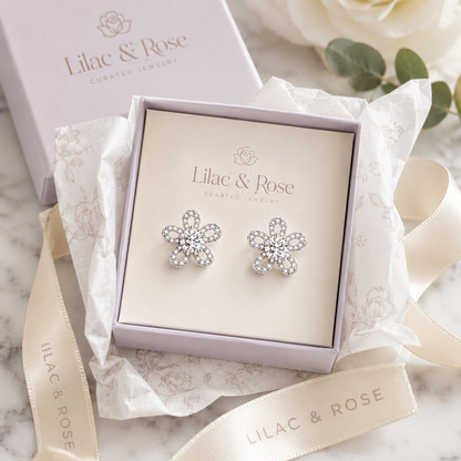 A pair of silver flower-shaped stud earrings set with clear crystals, presented in a lavender Lilac & Rose gift box with branded ribbon and packaging on a light marble surface.