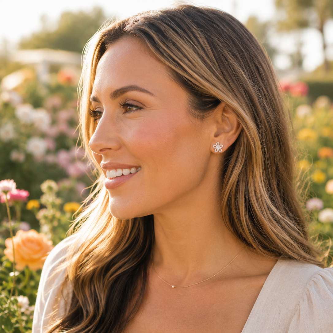 A woman with long highlighted brown hair is shown in profile outdoors in a sunlit flower garden, wearing a small silver stud earring and a delicate necklace. She is wearing a light beige top.