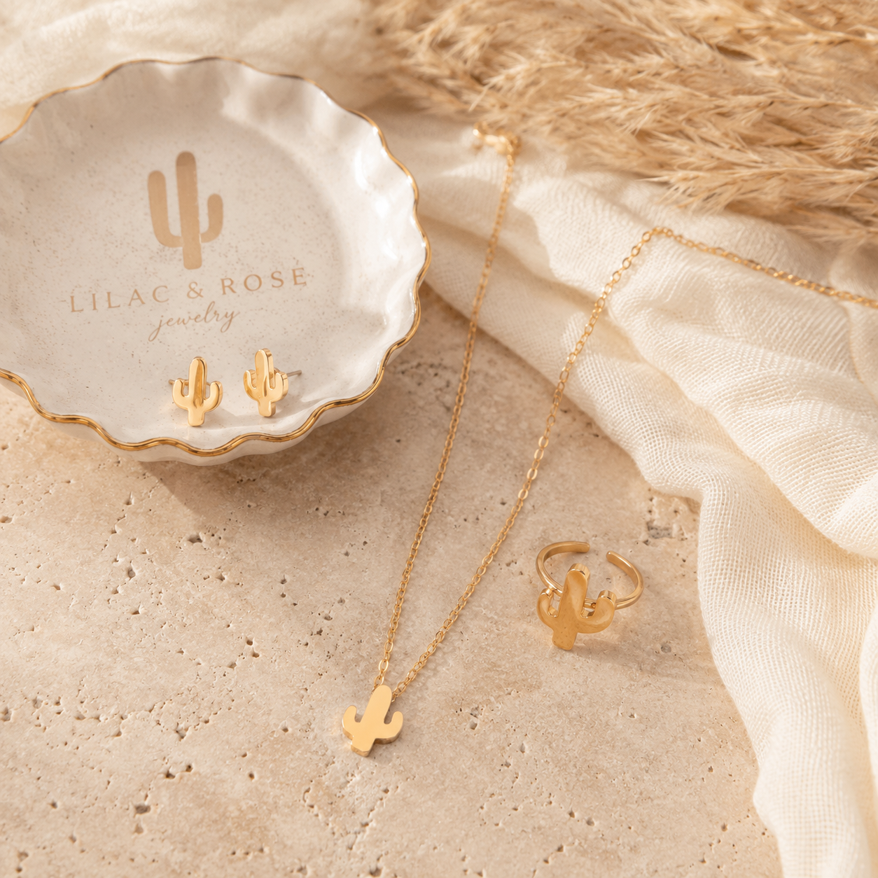 Gold jewelry set featuring a cactus pendant necklace, matching cactus stud earrings, and a cactus ring, styled on a beige stone surface with a white dish branded 'LILAC & ROSE jewelry' and cream fabric.