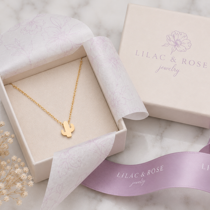 A gold necklace with a small cactus pendant displayed in an open cream jewelry box, with the branded closed box and lavender ribbon beside it on a light marble surface.