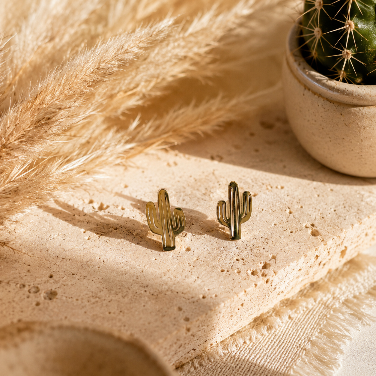 A pair of small Arizona cactus-shaped stud earrings in a gold-tone finish, displayed on a textured beige stone slab with dried pampas grass and a small potted cactus in the background.