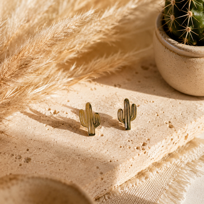 A pair of small Arizona cactus-shaped stud earrings in a gold-tone finish, displayed on a textured beige stone slab with dried pampas grass and a small potted cactus in the background.