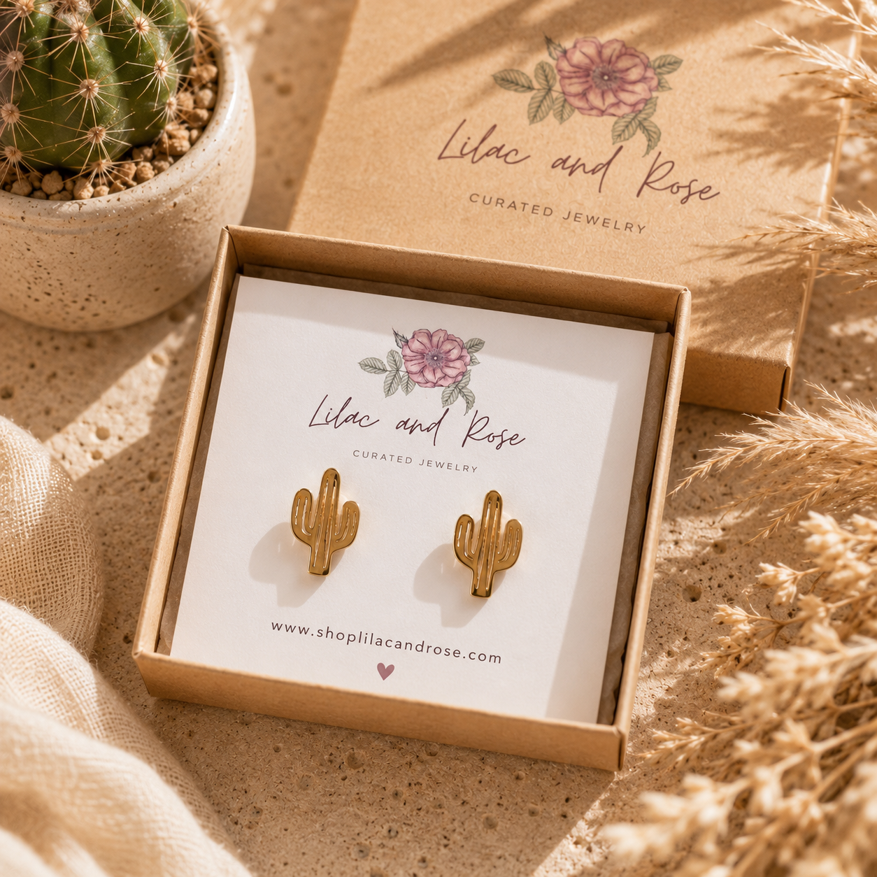 Gold cactus-shaped stud earrings displayed in a kraft jewelry box on a white insert card, with the matching branded box lid behind it and a small cactus plant nearby on a sandy surface.