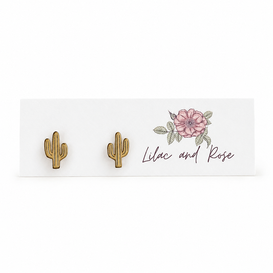 A pair of small gold cactus stud earrings displayed on a white card with a pink flower illustration and the script text 'Lilac and Rose'.