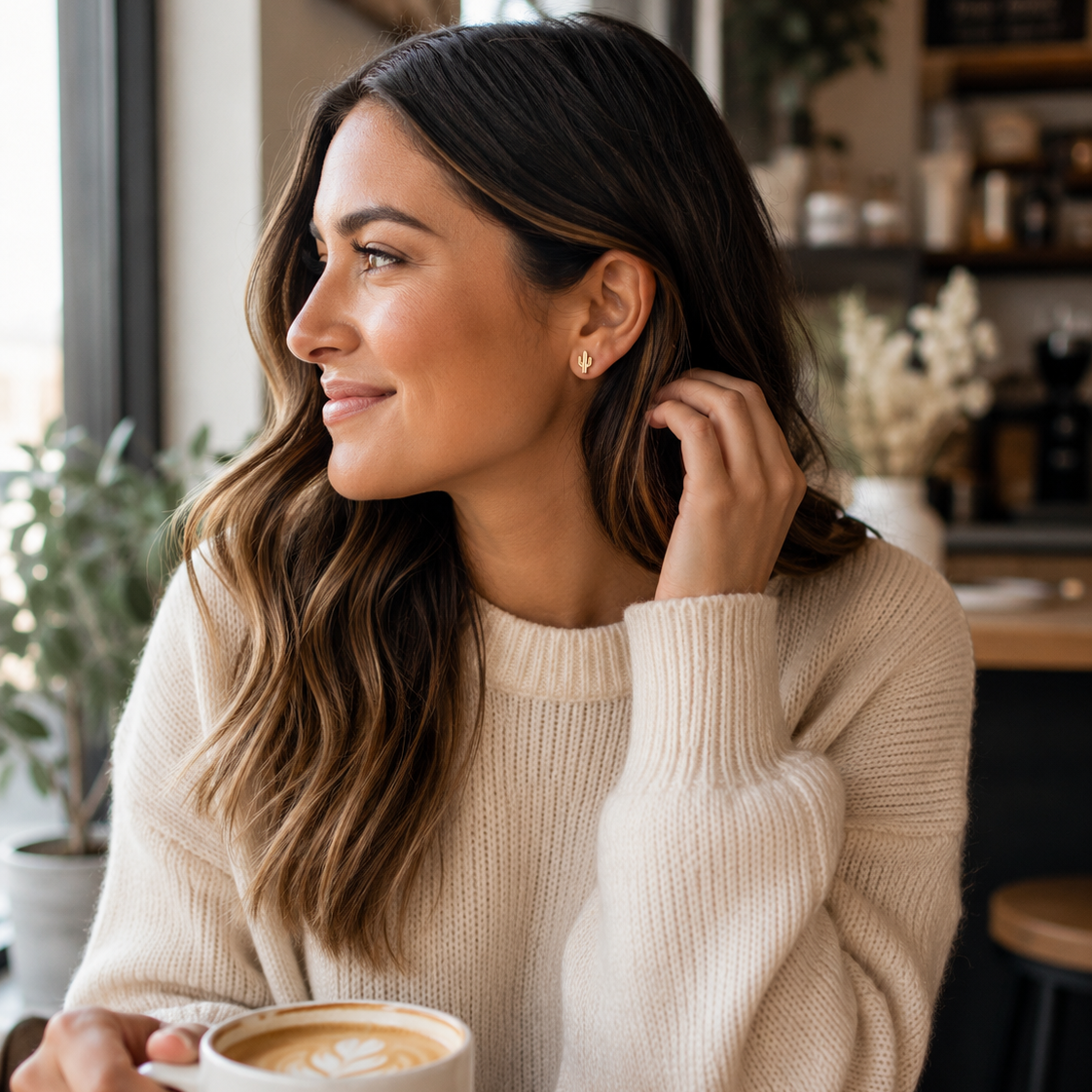 A woman with long wavy brown hair wearing a cream-colored ribbed knit sweater, sitting in a cozy cafe setting and holding a cup of latte art.