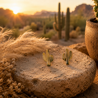 A pair of small gold cactus-shaped earrings displayed on a round stone slab in a desert setting at sunset, with cacti and dried grass in the background.