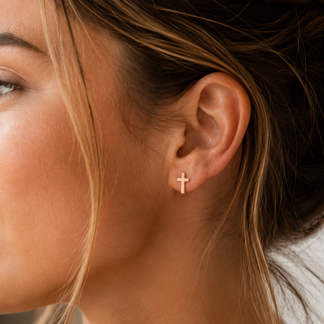 A close-up of a woman's ear wearing a small rose gold cross stud earring.