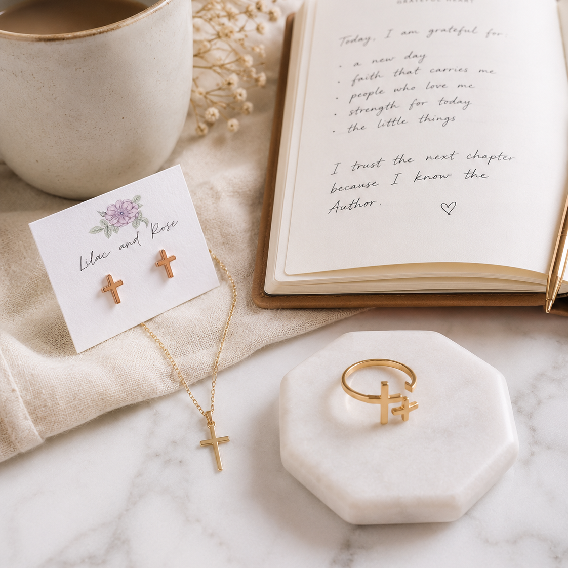 A styled flat-lay of gold cross jewelry on a marble surface, including a necklace with a cross pendant, a ring with a cross accent, and a pair of small cross stud earrings on a card. An open book, a ceramic mug, and dried flowers surround the jewelry.