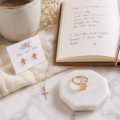 A styled flat-lay of gold cross jewelry on a marble surface, including a necklace with a cross pendant, a ring with a cross accent, and a pair of small cross stud earrings on a card. An open book, a ceramic mug, and dried flowers surround the jewelry.
