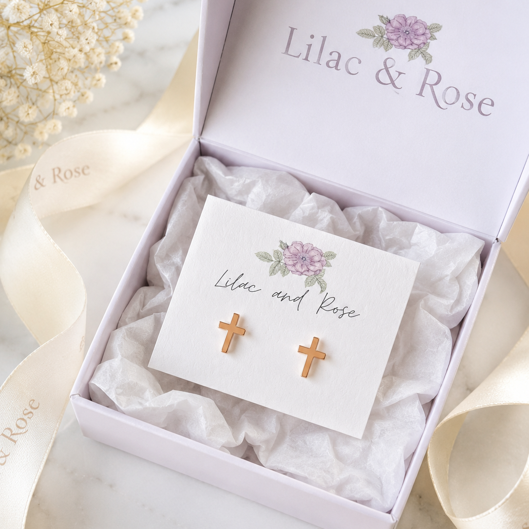 A pair of small gold cross stud earrings displayed on a white card inside an open white gift box with tissue paper. The box lid and ribbon are printed with the brand name 'Lilac & Rose', and dried flowers sit in the background on a light marble surface.