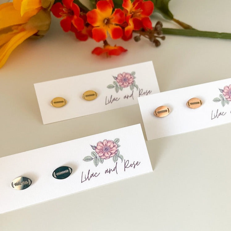 Stainless steel football stud earrings in silver and rose gold colors, displayed on a white card with a floral design and the words 'Lilac and Rose' written on it.