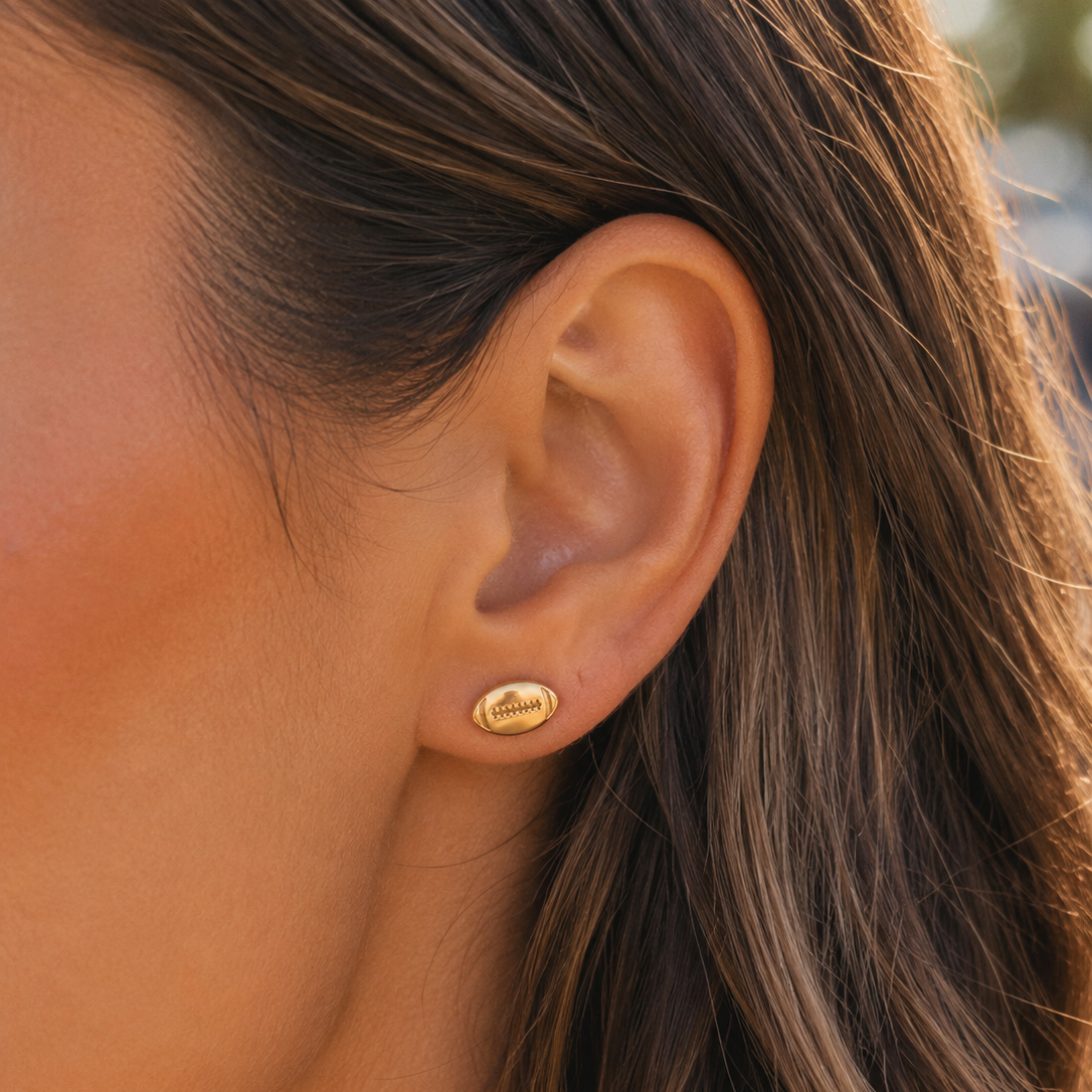 A close-up of a woman's ear wearing a small football gold stud earring.