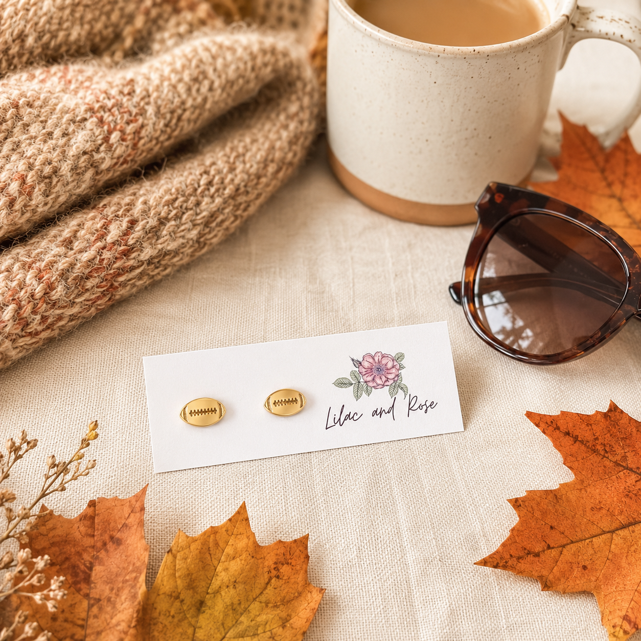 Two small gold stud earrings displayed on a white card with a floral logo reading 'Lilac and Rose,' styled on a beige fabric surface with autumn leaves, a mug, sunglasses, and a knitted scarf.